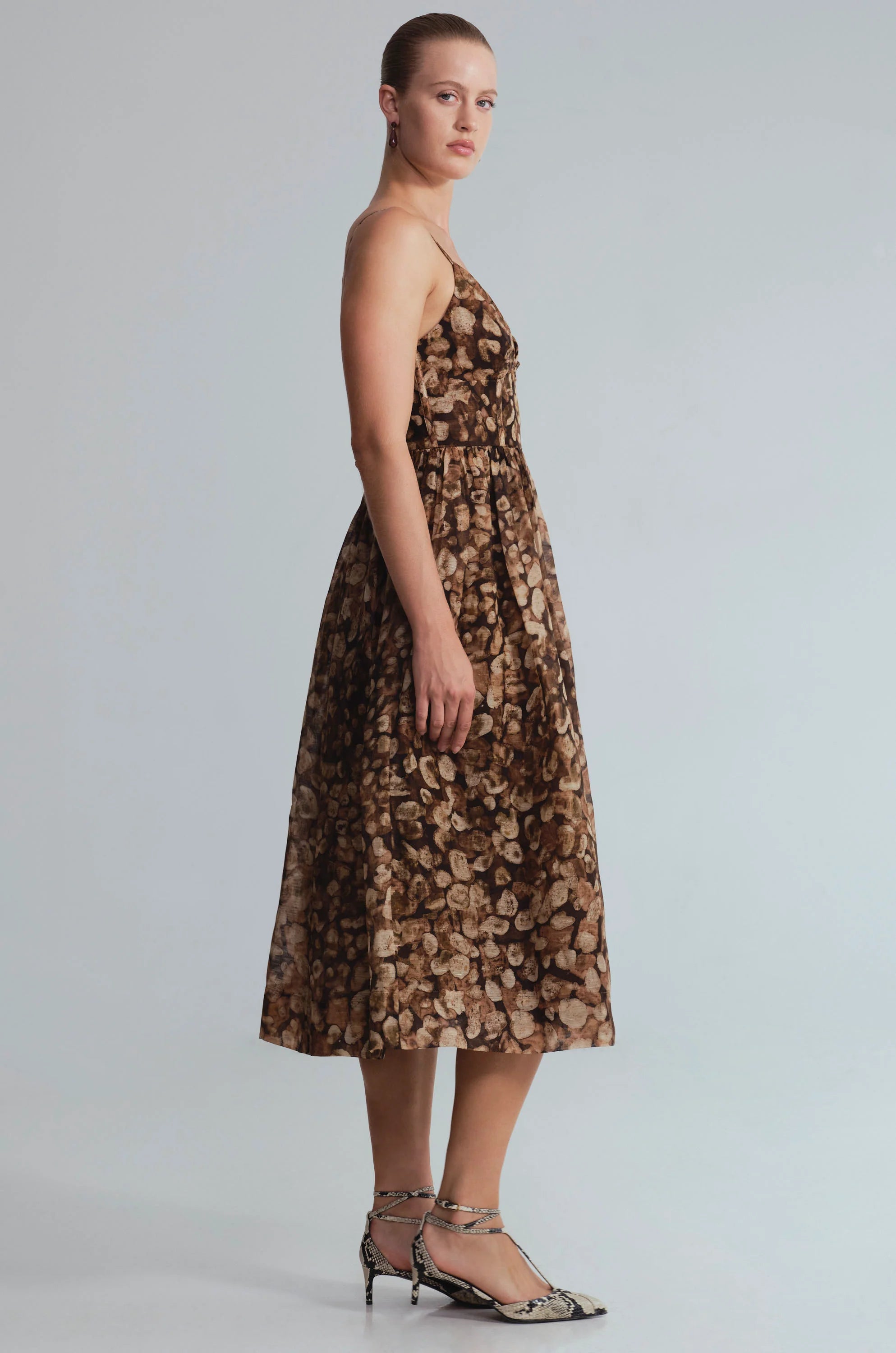 A woman models a sleeveless brown midi dress with a beige and gold abstract floral pattern, paired with metallic silver strappy heels, against a light gray studio background.