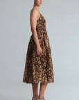 A woman models a sleeveless brown midi dress with a beige and gold abstract floral pattern, paired with metallic silver strappy heels, against a light gray studio background.
