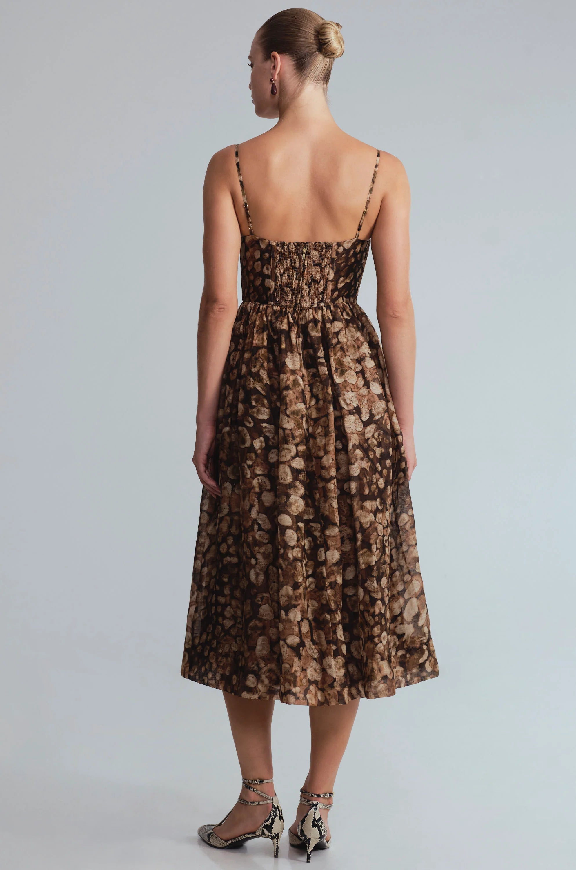 A woman viewed from the back wearing a sleeveless midi dress with thin straps, a smocked bodice, and a brown and beige floral pattern. She is standing against a light gray studio background.