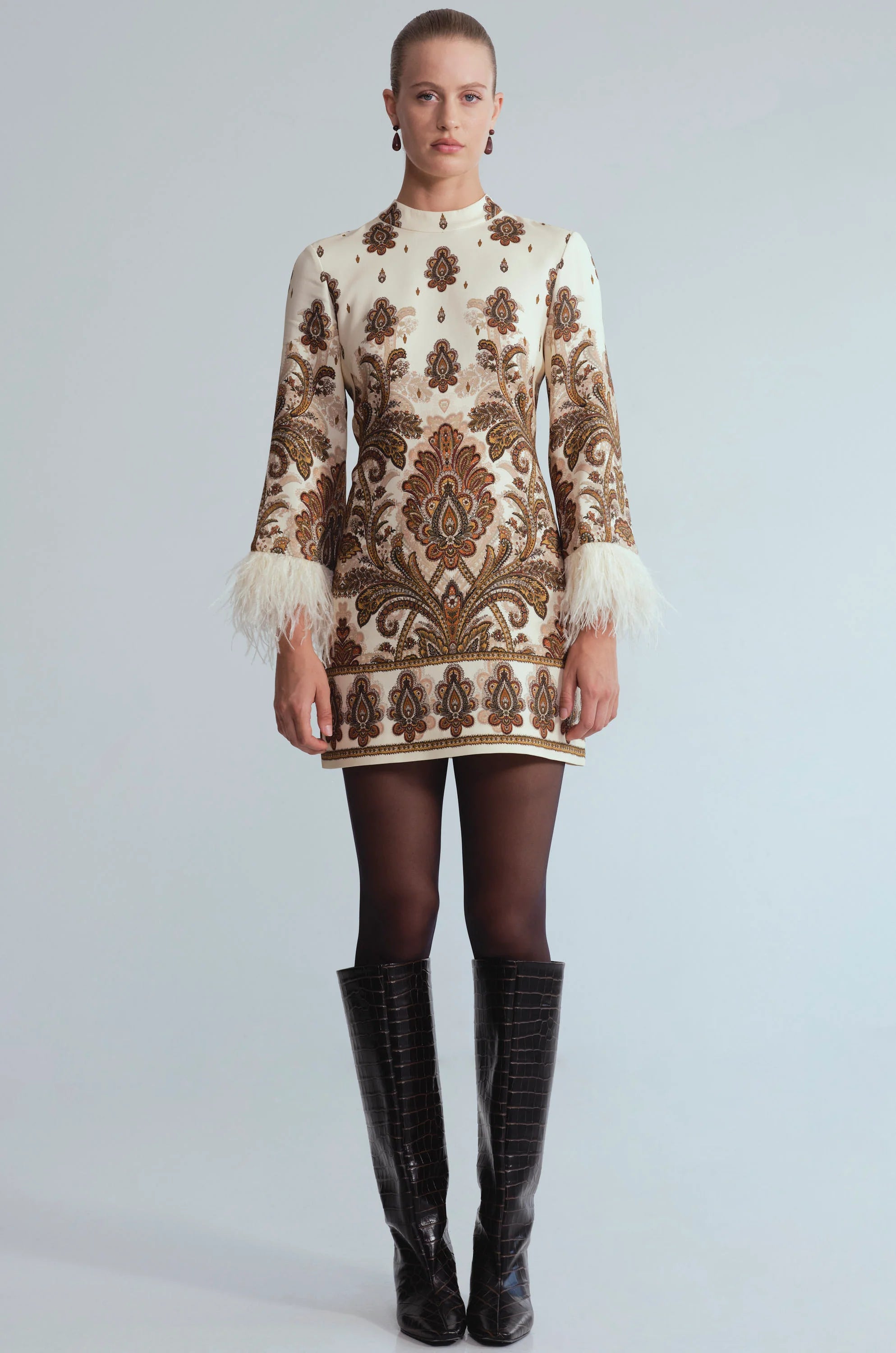 A model stands against a light gray studio background wearing a cream and brown ornate patterned mini dress with feathered cuffs, sheer black tights, and black knee-high boots.