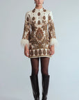 A model stands against a light gray studio background wearing a cream and brown ornate patterned mini dress with feathered cuffs, sheer black tights, and black knee-high boots.