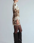 A model stands against a light gray studio background wearing a cream and brown ornate patterned mini dress with feathered cuffs, sheer black tights, and black knee-high boots, side