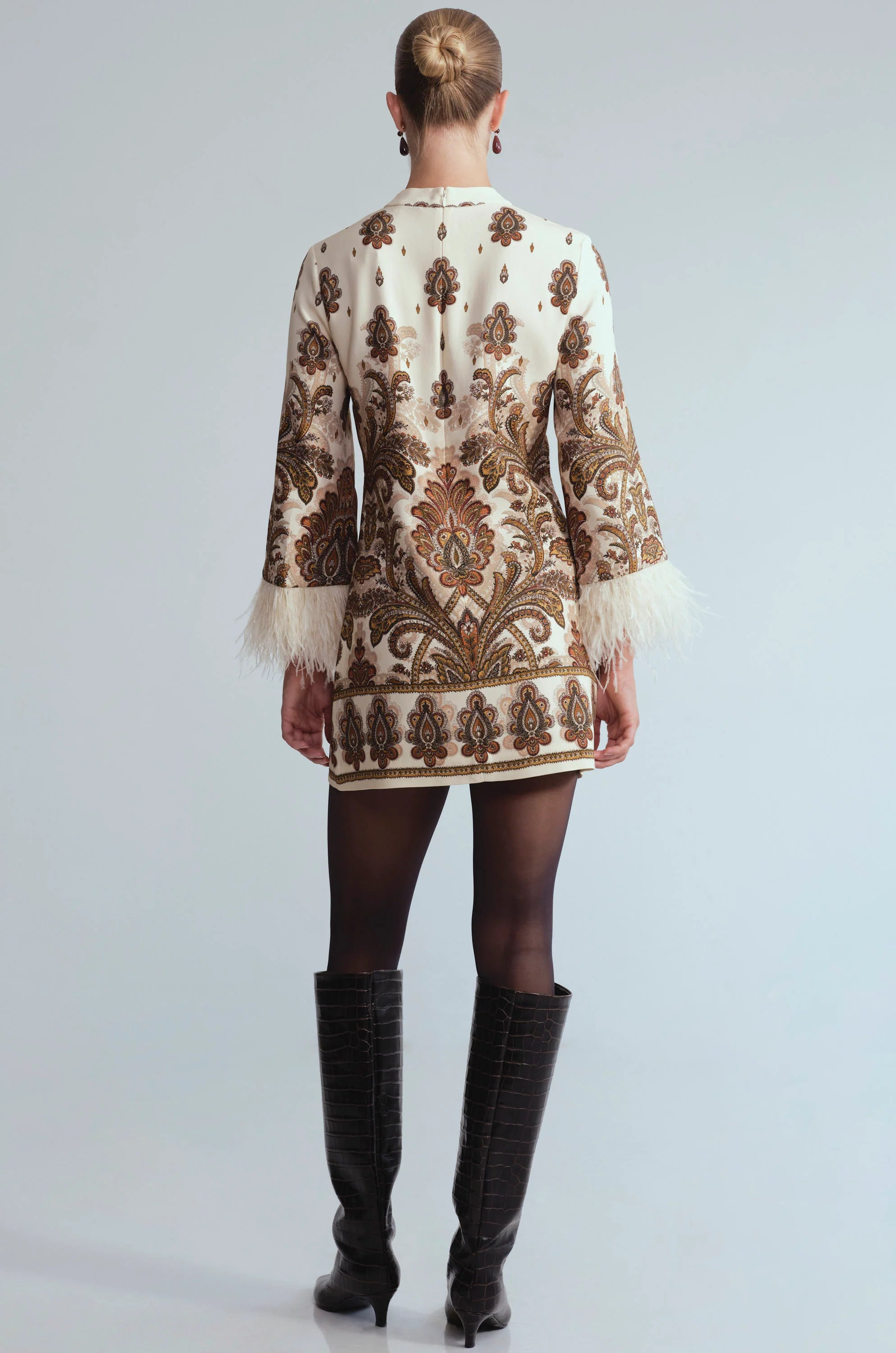 A model shown from the back wearing a short cream-colored jacket with intricate brown and gold paisley embroidery, white feather trim at the cuffs, black tights, and black knee-high boots, set against a light gray studio background.
