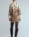 A model shown from the back wearing a short cream-colored jacket with intricate brown and gold paisley embroidery, white feather trim at the cuffs, black tights, and black knee-high boots, set against a light gray studio background.