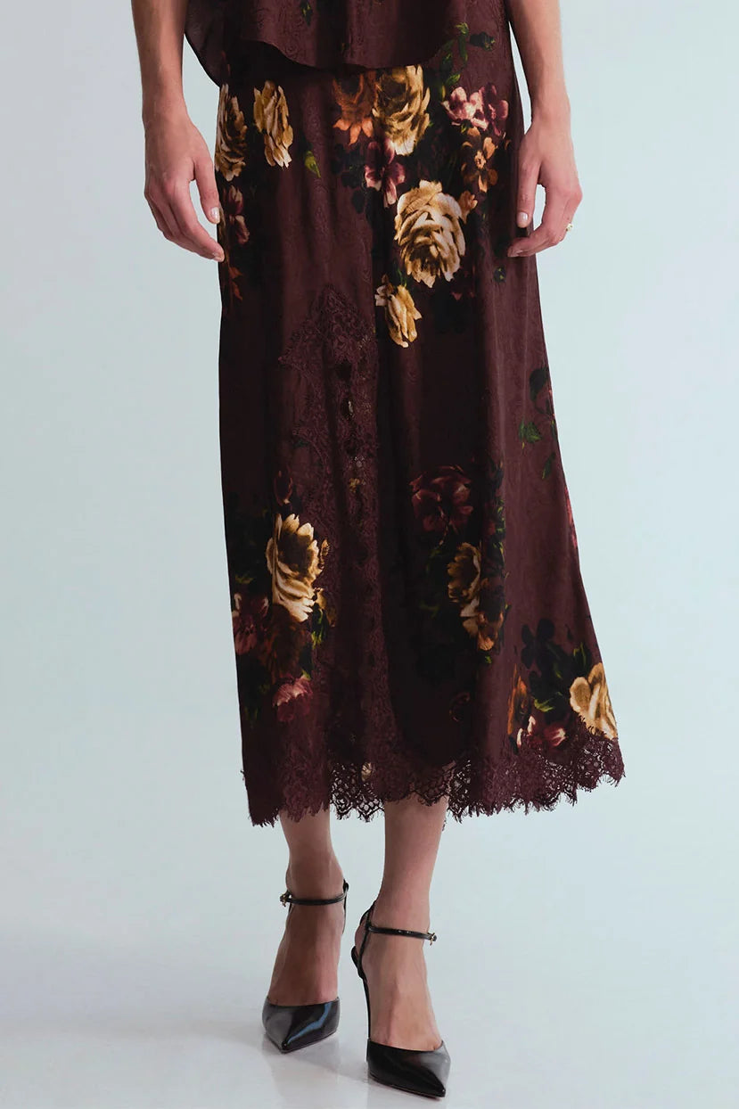 A dark brown midi skirt with a large floral print in beige, gold, and red, featuring a lace hem, shown on a model wearing black pointed-toe heels against a light gray background.