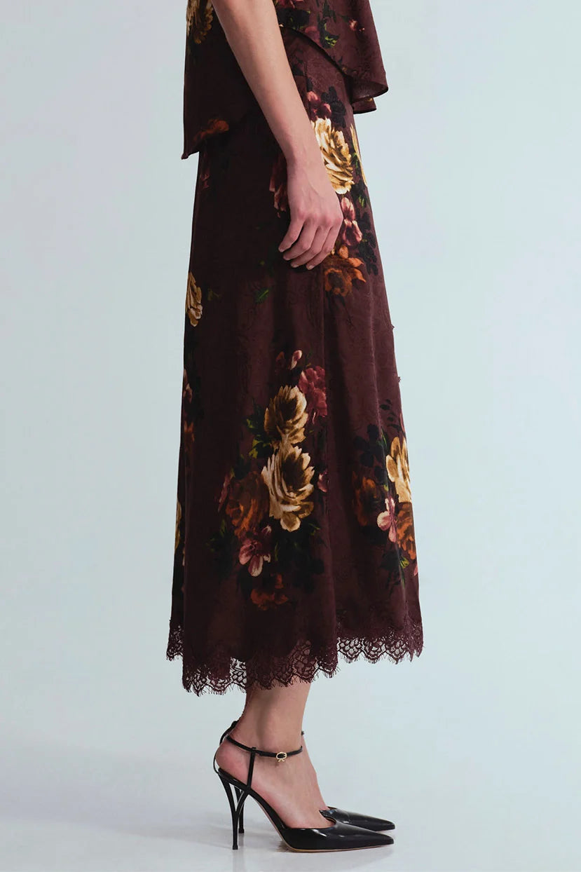 A model wearing a dark brown midi skirt with a gold and orange floral print and a lace hem, paired with black high heels, against a light gray background.