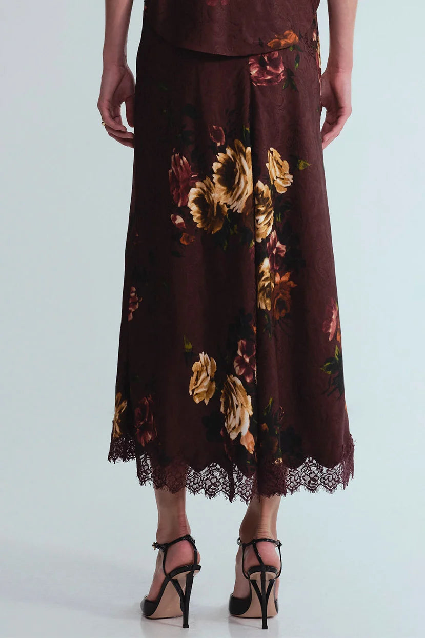 A dark brown midi skirt with a large floral print in gold, beige, and muted red, featuring a lace hem and worn with black strappy heels against a light gray background.