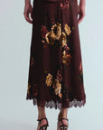 A dark brown midi skirt with a large floral print in gold, beige, and muted red, featuring a lace hem and worn with black strappy heels against a light gray background.