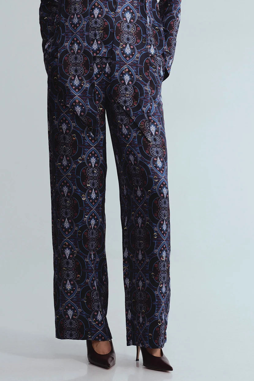 A person wearing dark navy patterned trousers with an ornate medallion print in blue, gold, and white, paired with dark brown pointed-toe heels against a light gray studio background.