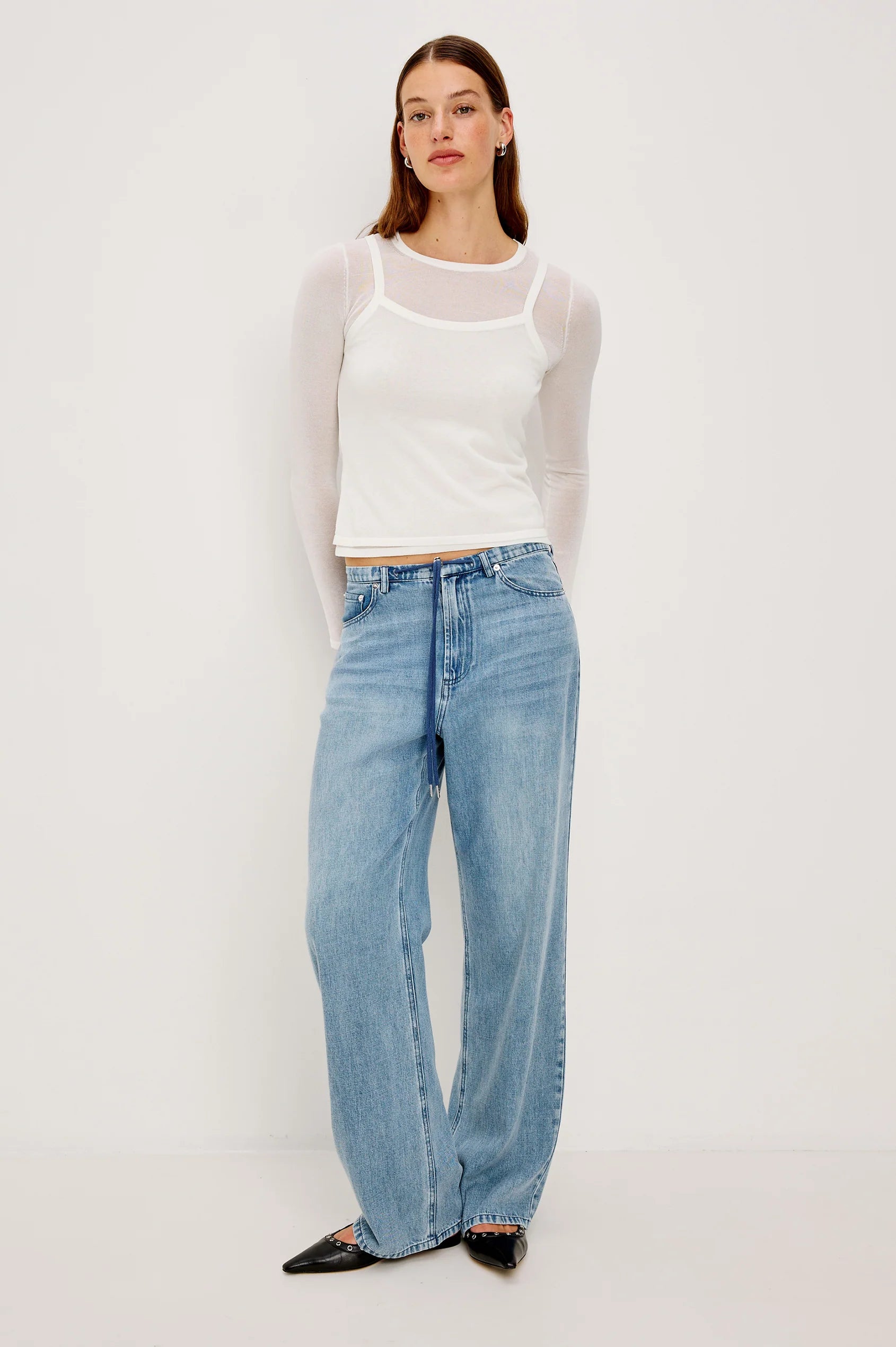 Woman wearing a white tank top and light blue jeans on a plain background front view