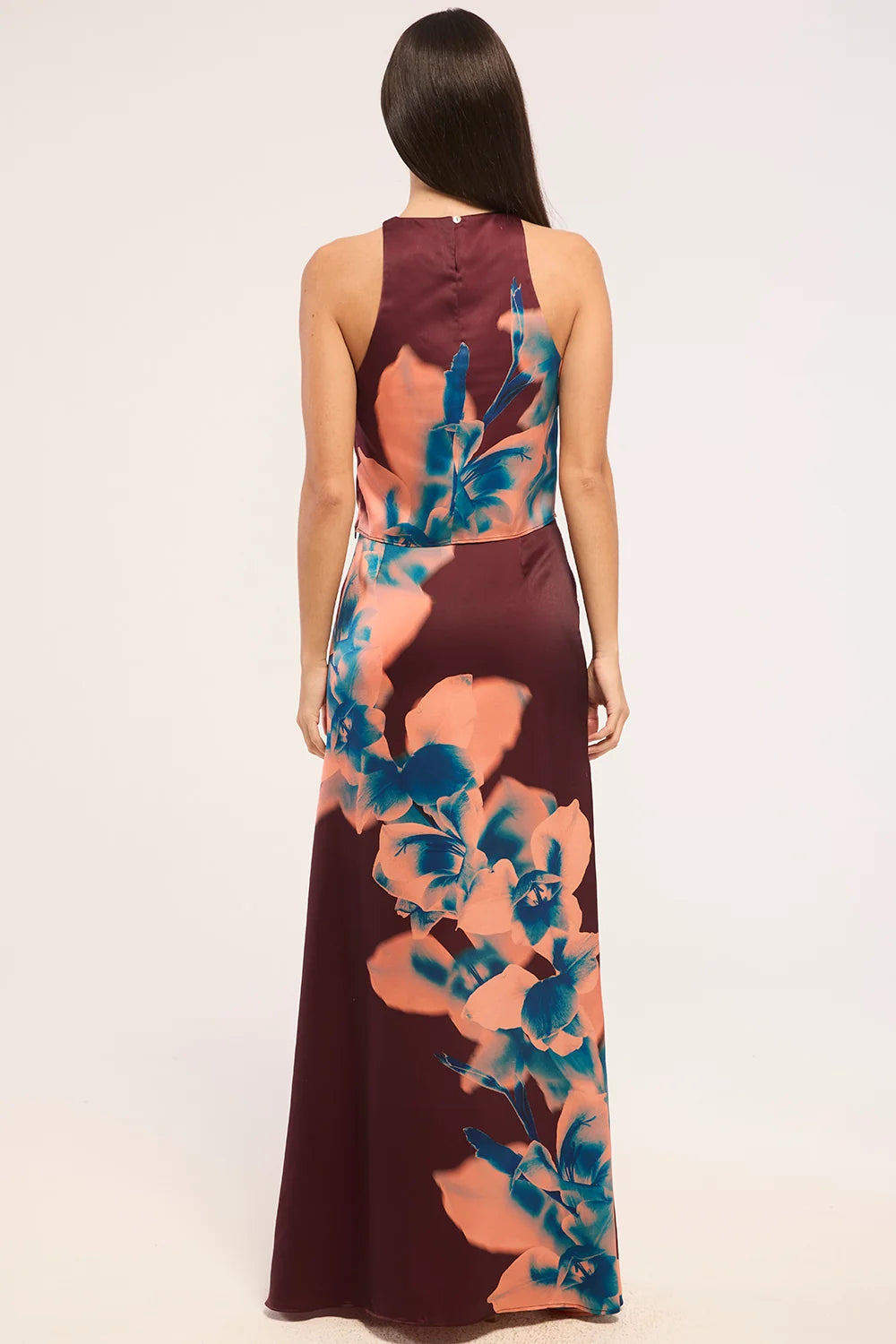 A woman viewed from the back wearing a sleeveless floor-length dress with a deep V-neckline and a large floral print in teal blue and peach over a dark burgundy base, against a plain light gray studio background.