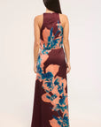 A woman viewed from the back wearing a sleeveless floor-length dress with a deep V-neckline and a large floral print in teal blue and peach over a dark burgundy base, against a plain light gray studio background.