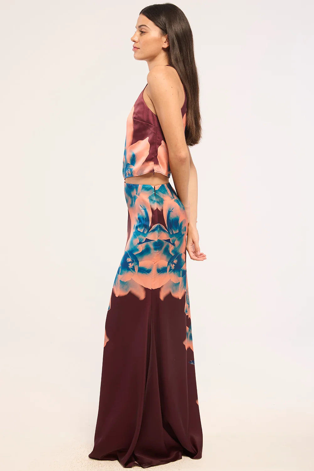 A woman modeling a sleeveless two-piece set with a halter crop top and matching high-waisted skirt, featuring a multicolor abstract print in teal, peach, and deep burgundy on a plain light background.