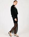 A woman modeling a black long-sleeve blouse with a high neck and pleated details, paired with a matching black ruched skirt and sheer black lace tights, against a plain light gray studio background.