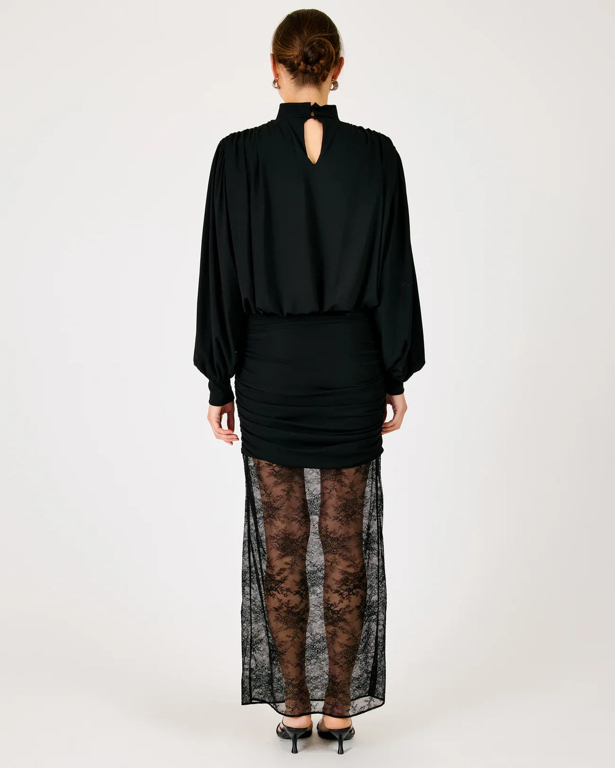 A model viewed from the back wearing a black long-sleeve top with a keyhole cutout, a ruched black mini skirt, sheer black lace tights, and black heels, set against a plain light gray studio background.