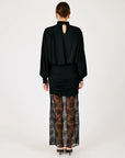 A model viewed from the back wearing a black long-sleeve top with a keyhole cutout, a ruched black mini skirt, sheer black lace tights, and black heels, set against a plain light gray studio background.