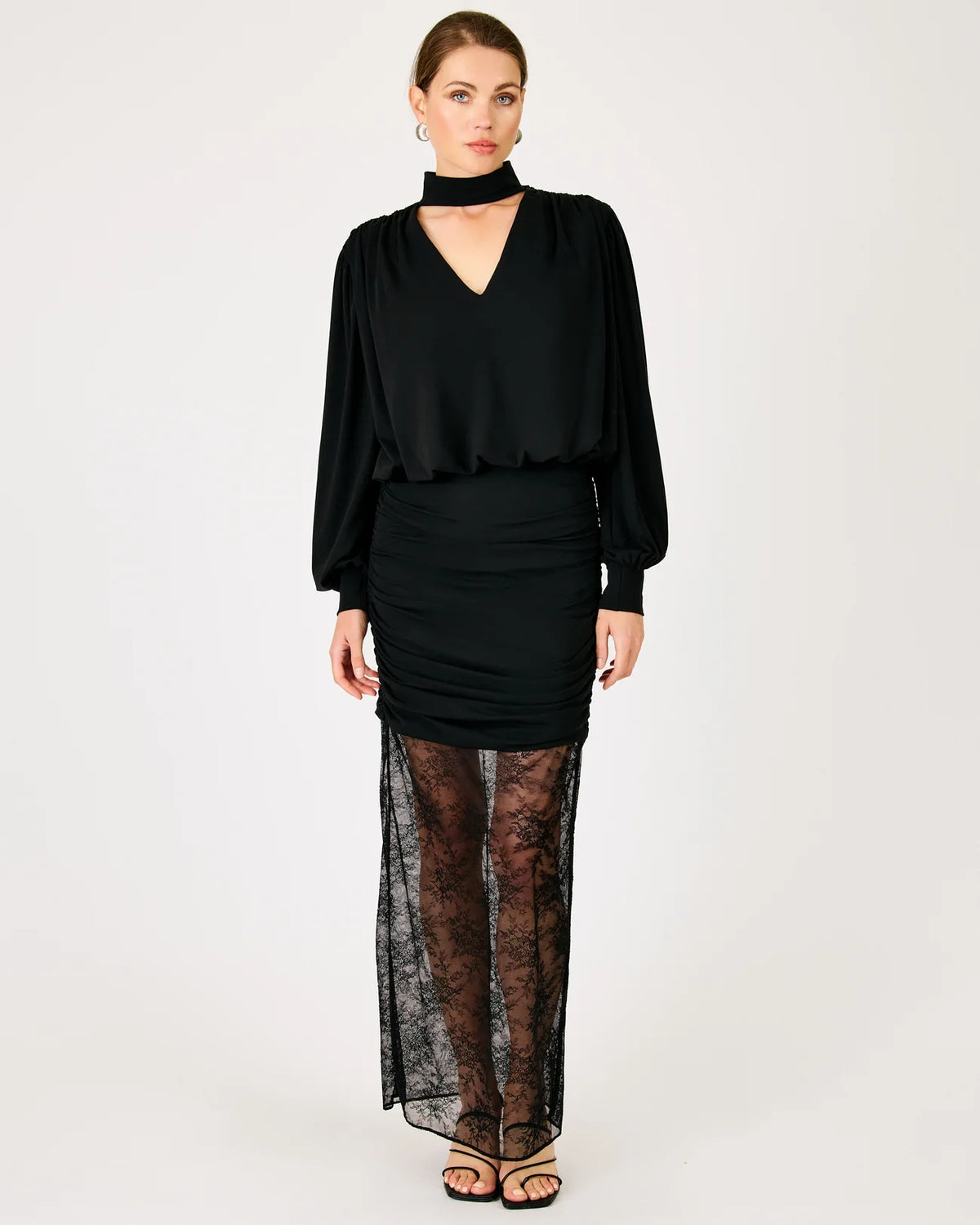A model wearing a black long-sleeve mock-neck top with a draped waist, paired with a ruched black mini skirt and sheer black lace tights, standing against a plain white studio background.