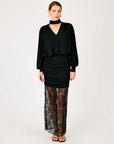 A model wearing a black long-sleeve mock-neck top with a draped waist, paired with a ruched black mini skirt and sheer black lace tights, standing against a plain white studio background.