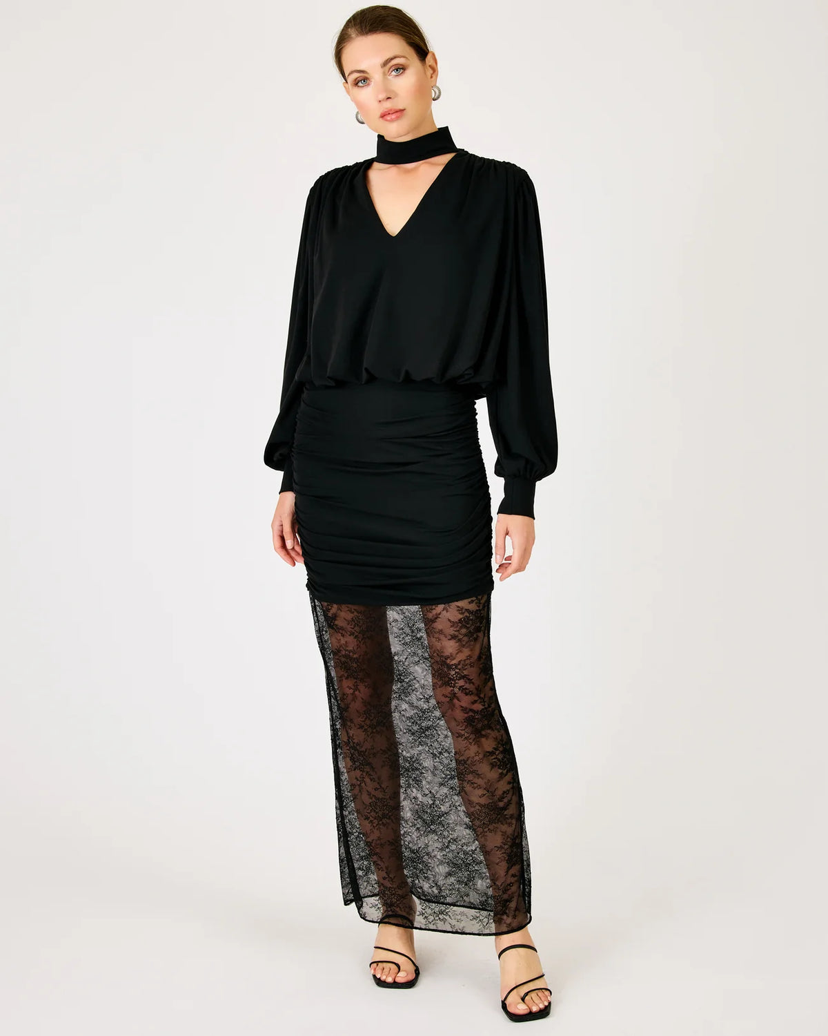 A model wearing a black long-sleeve blouse with a high neck, a ruched black mini skirt, and sheer black lace tights, paired with black strappy heels, against a plain light gray studio background.