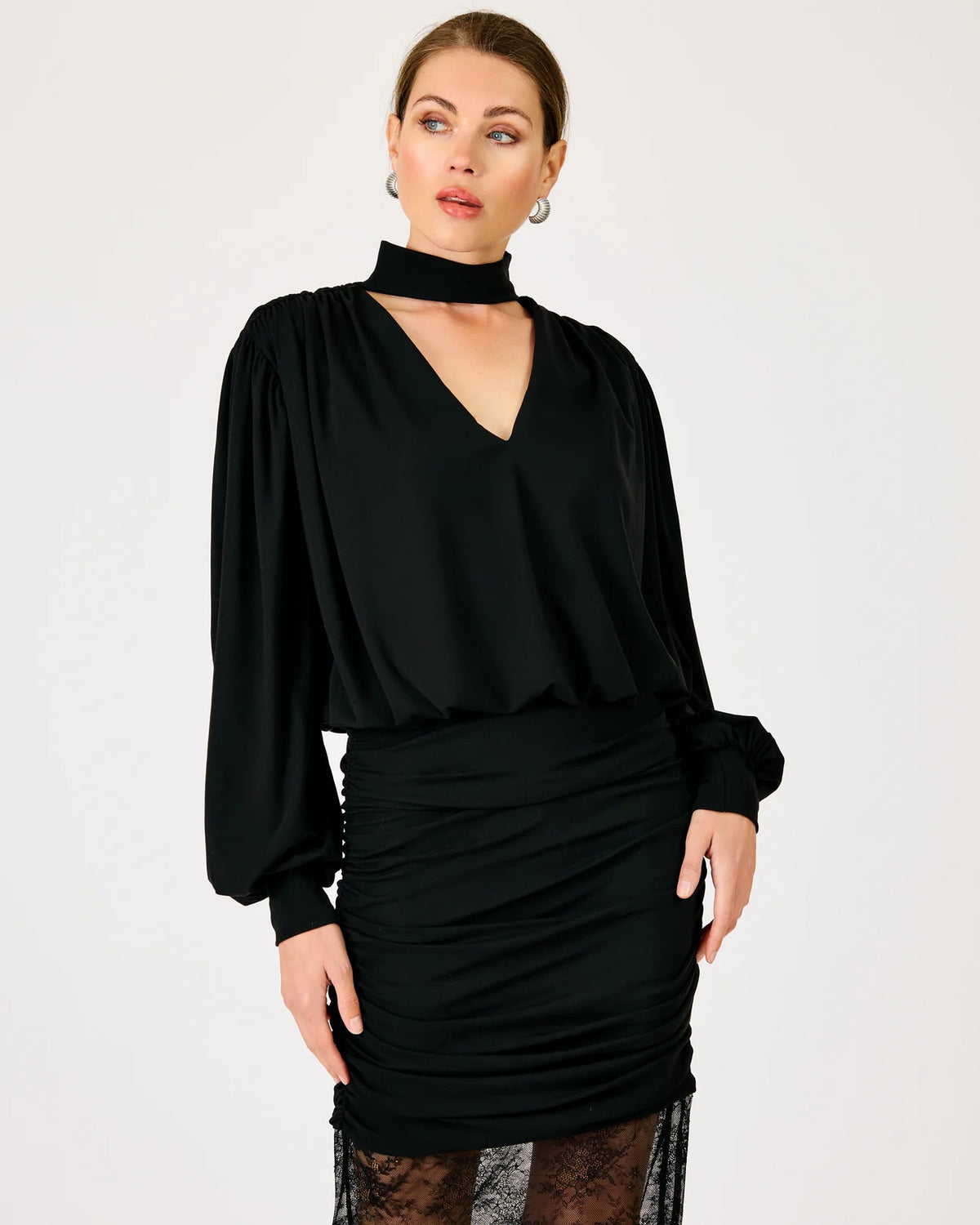 A woman modeling a black long-sleeve blouse with a high mock neck and a ruched black skirt, photographed against a plain light gray studio background.