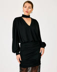 A woman modeling a black long-sleeve blouse with a high mock neck and a ruched black skirt, photographed against a plain light gray studio background.