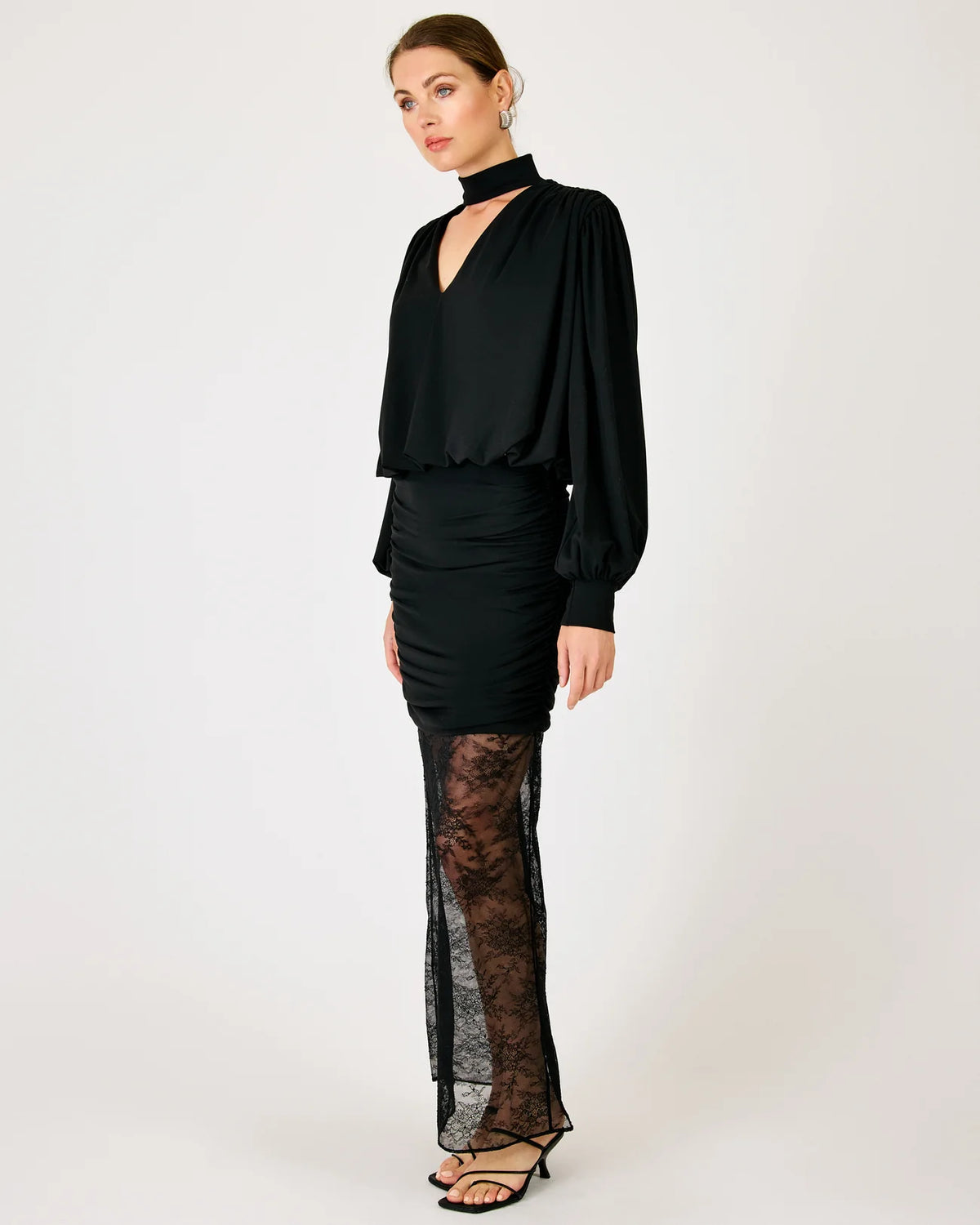 A model wearing a black long-sleeve blouse with a high neck, a ruched black mini skirt, and sheer black lace tights, standing against a plain light gray studio background.