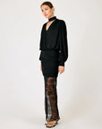 A model wearing a black long-sleeve blouse with a high neck, a ruched black mini skirt, and sheer black lace tights, standing against a plain light gray studio background.