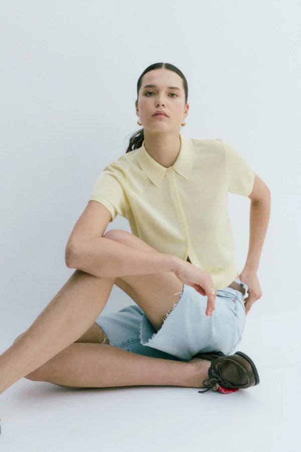 Woman sitting on a white floor wearing a light yellow shirt and denim shorts.