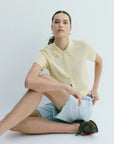Woman sitting on a white floor wearing a light yellow shirt and denim shorts.