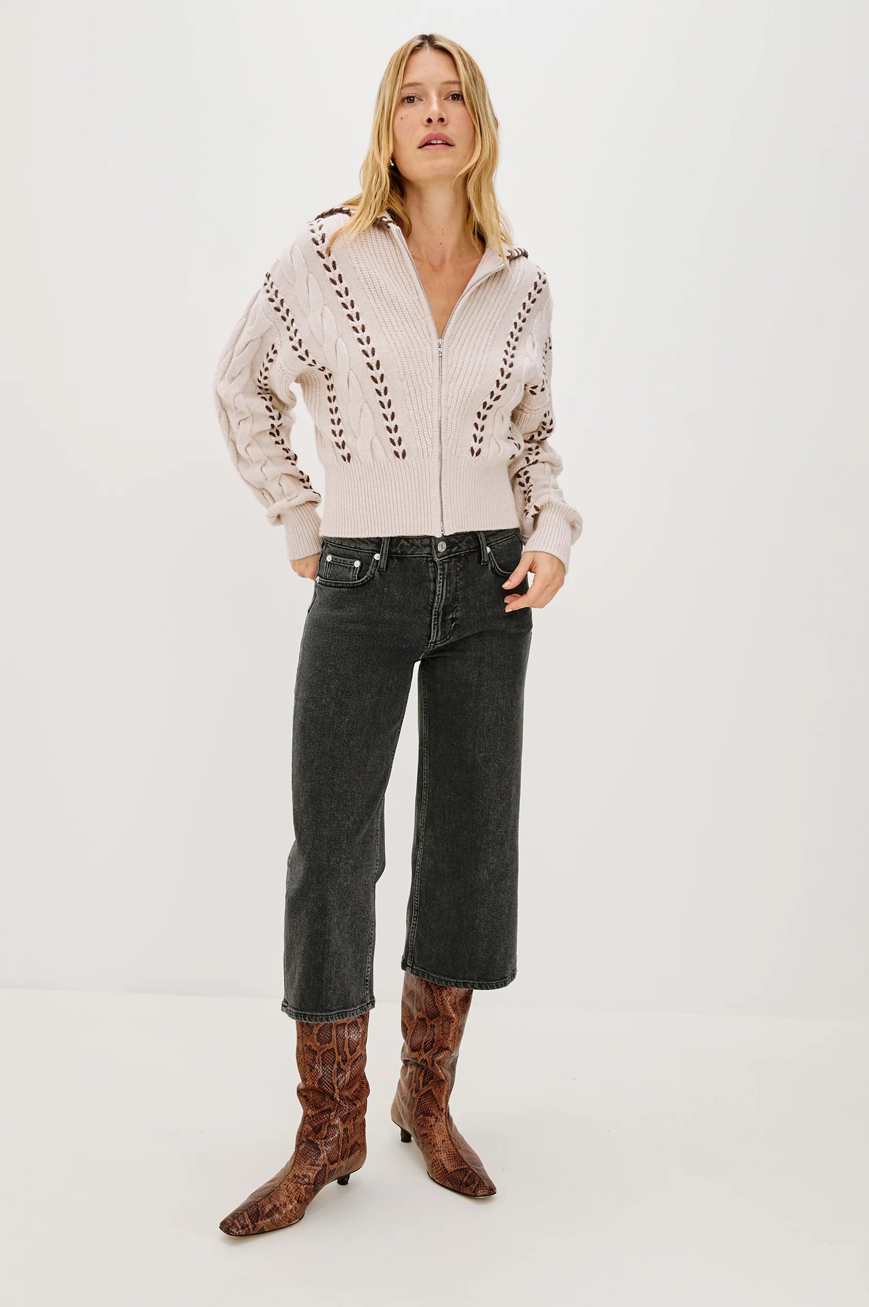 Woman wearing a beige zip-up sweater with lace details, black jeans, and brown boots on a white background