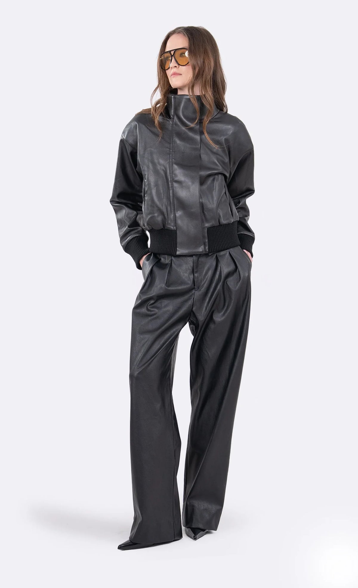 A model wearing a black faux-leather jumpsuit with a high collar, ribbed waistband, and wide-leg pants, styled with gold-framed sunglasses against a light gray background.