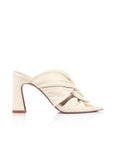 A single cream-colored leather mule sandal with a chunky block heel, featuring a criss-cross strap design and a metallic bronze accent band, shown from a side profile on a white background.