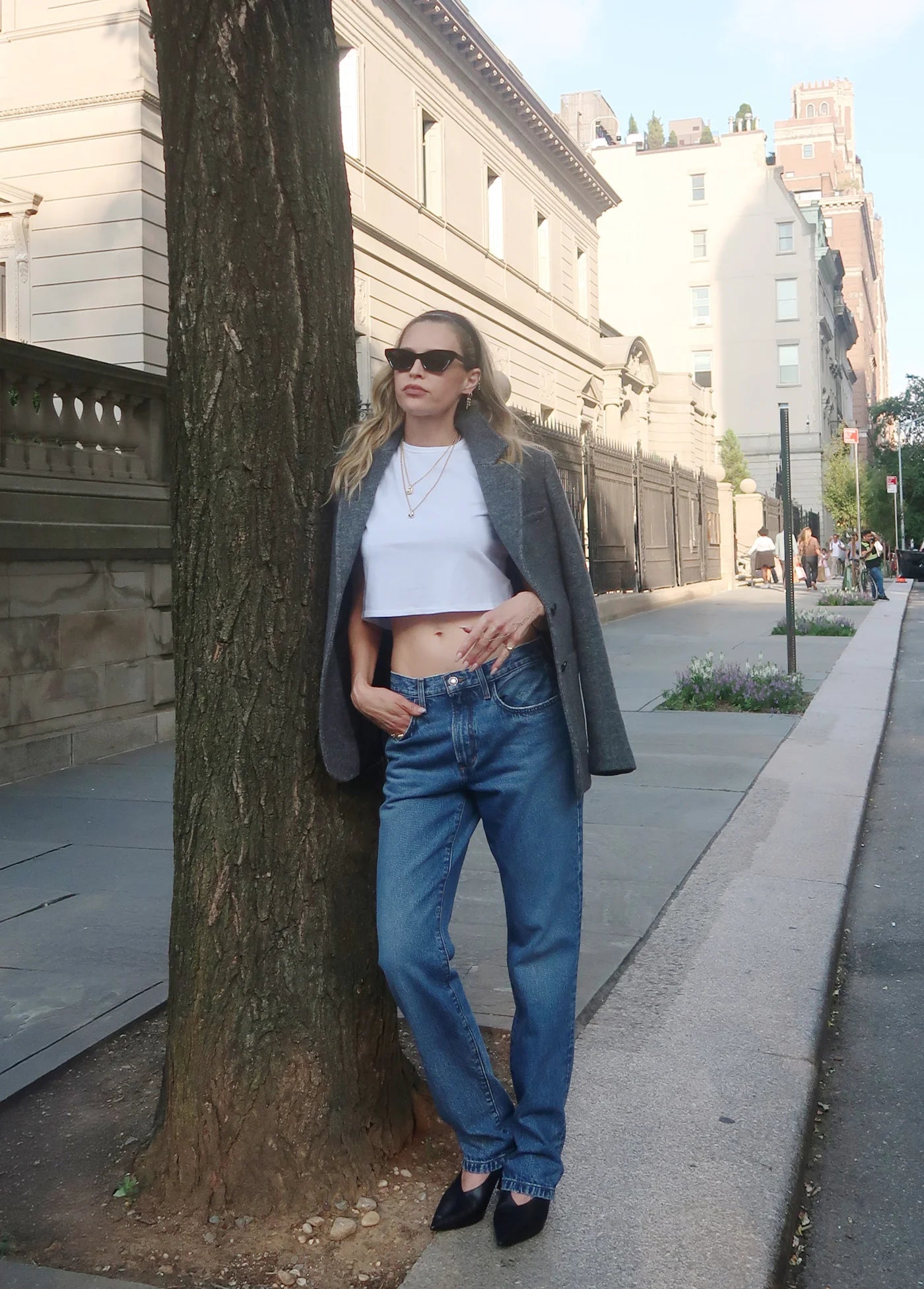 Woman standing against a tree on a city sidewalk wearing sunglasses, a white crop top, blue jeans, and a gray coat.