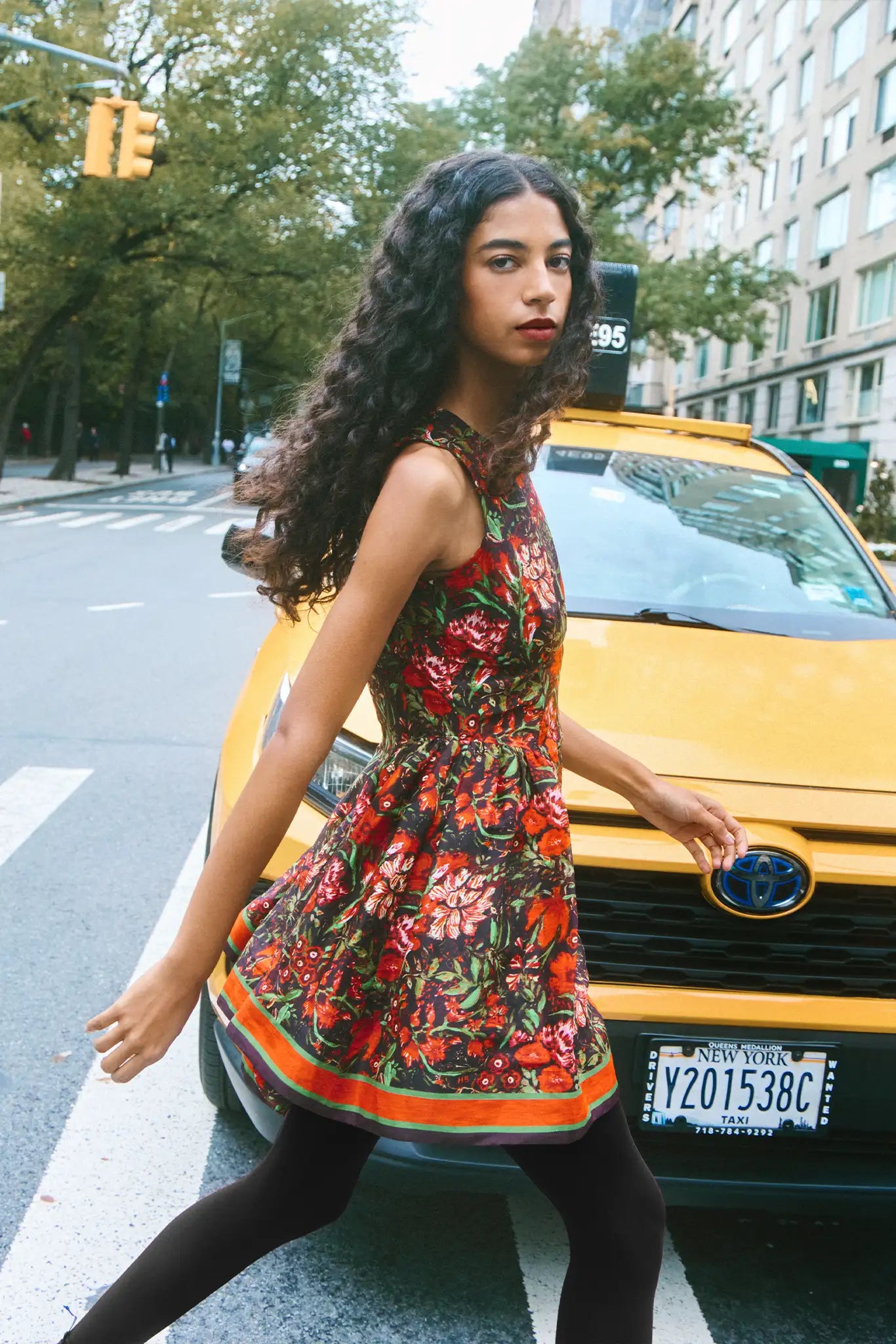 Woman in a floral dress crossing a street with a yellow taxi in the background