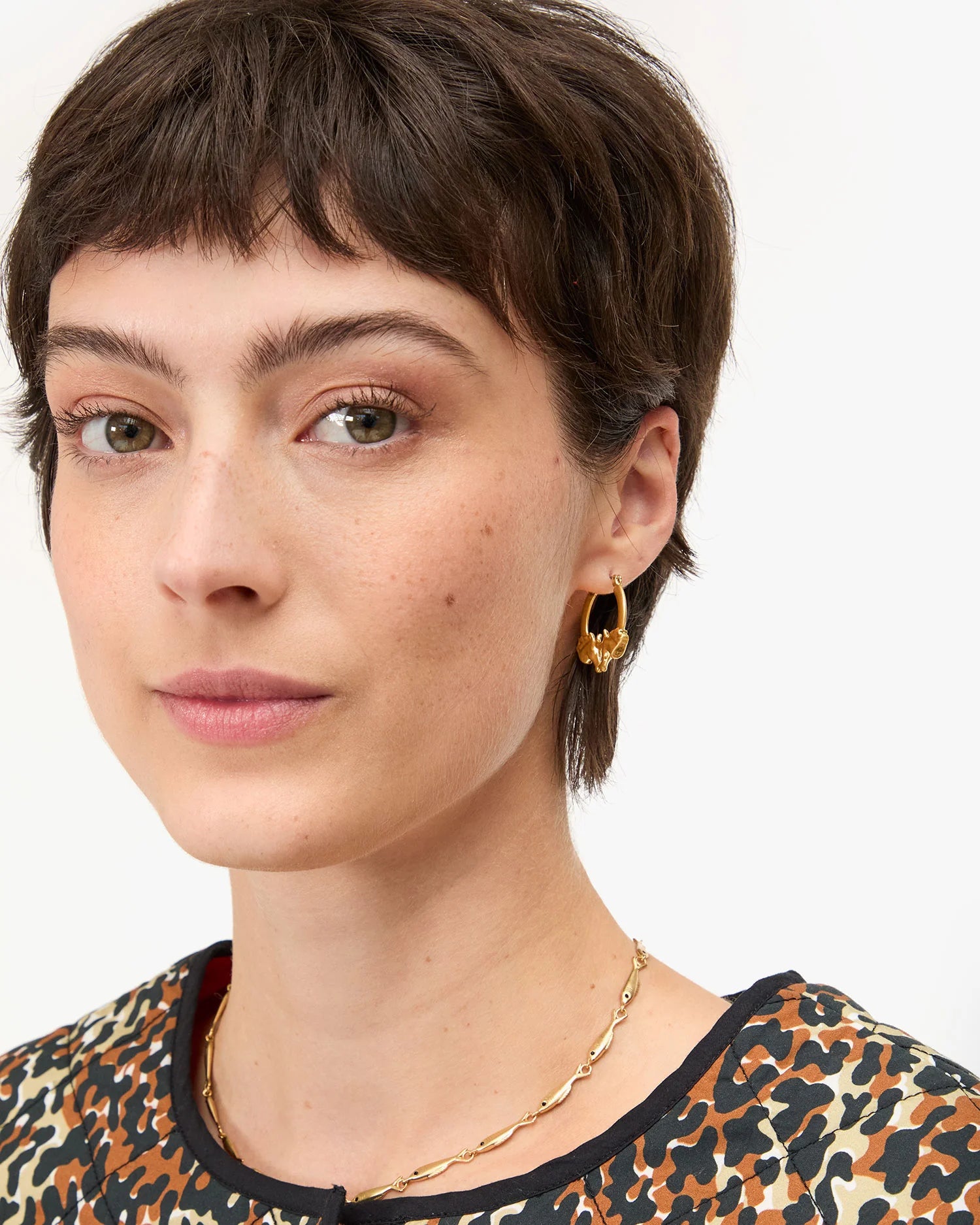 Close-up of a person with short hair wearing gold earrings and a necklace on a white background
