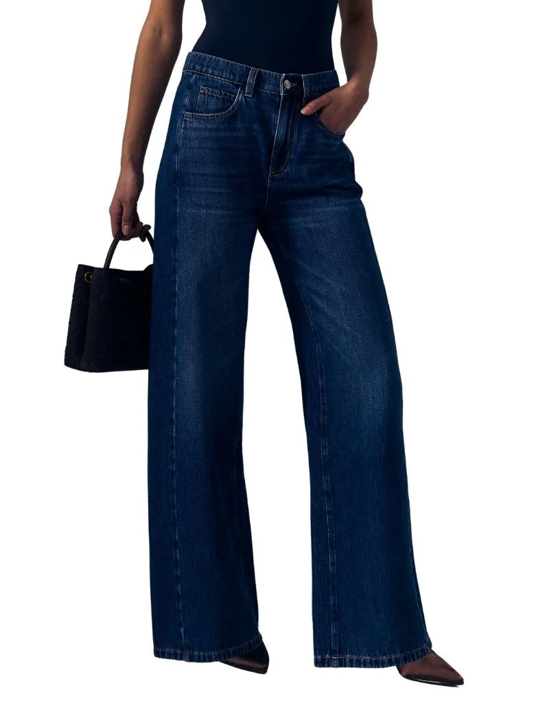 A model wearing dark blue wide-leg jeans, holding a black handbag, against a plain white background.
