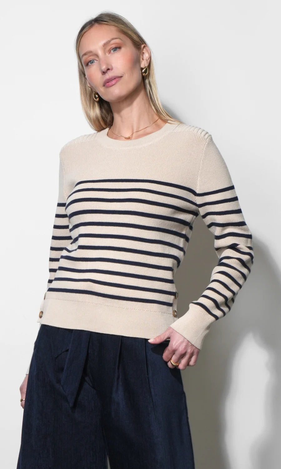A woman wearing a cream and navy blue horizontally striped knit sweater with dark navy wide-leg trousers, standing against a plain light gray wall.