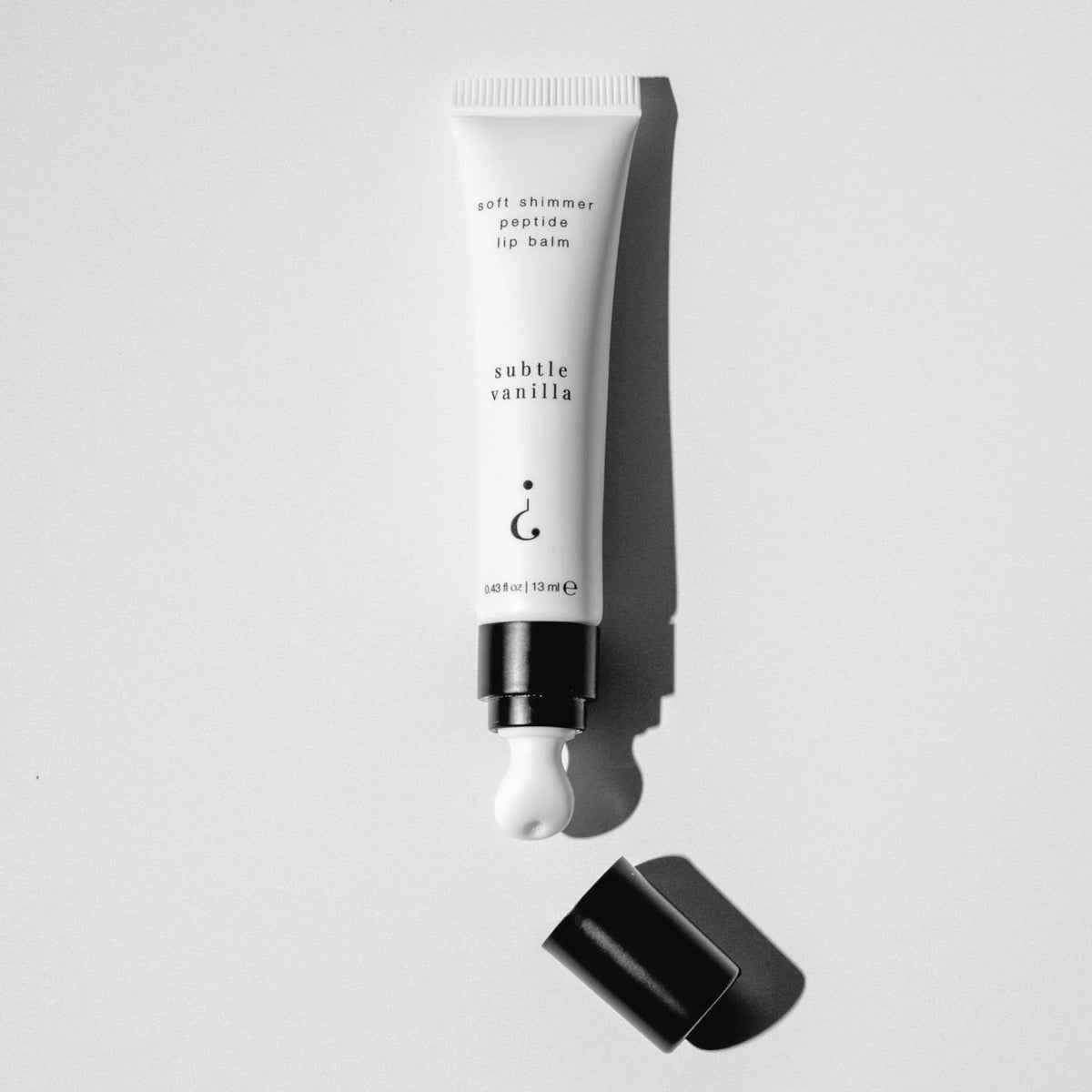 A white cosmetic tube of lip balm with black text reading 'soft shimmer peptide lip balm' and 'subtle vanilla,' shown uncapped with black cap on a light gray background.