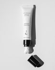 A white cosmetic tube of lip balm with black text reading 'soft shimmer peptide lip balm' and 'subtle vanilla,' shown uncapped with black cap on a light gray background.