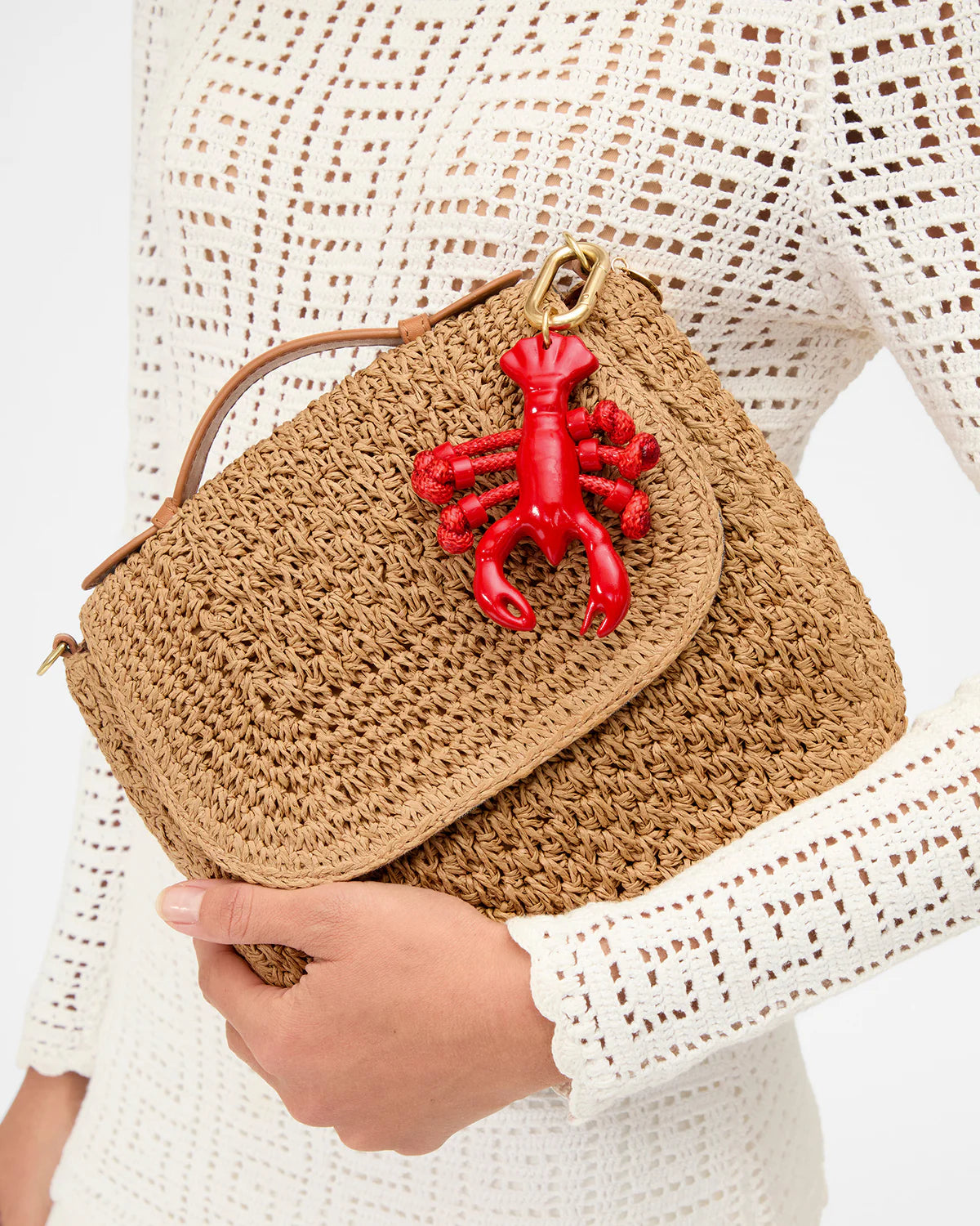 Brown woven handbag with a red lobster charm held by a person wearing a white lace top.