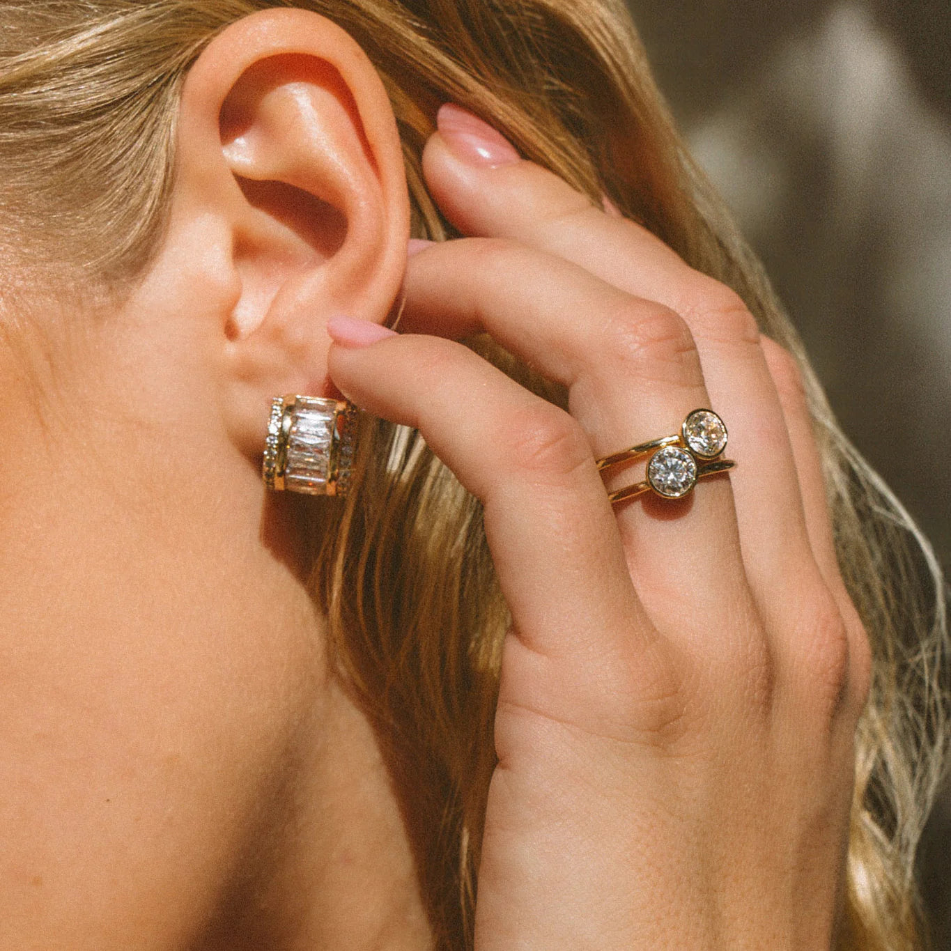 Close-up of a person wearing diamond earrings and a ring, with a blurred background.