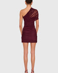 A woman viewed from the back wearing a ruched one-shoulder mini dress in deep burgundy, paired with dark heeled sandals, against a light gray studio background.