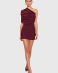 A woman modeling a ruched, one-shoulder mini dress in deep burgundy against a solid light gray background.