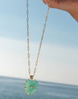 Necklace with a heart-shaped green pendant held against a blue sky and ocean background
