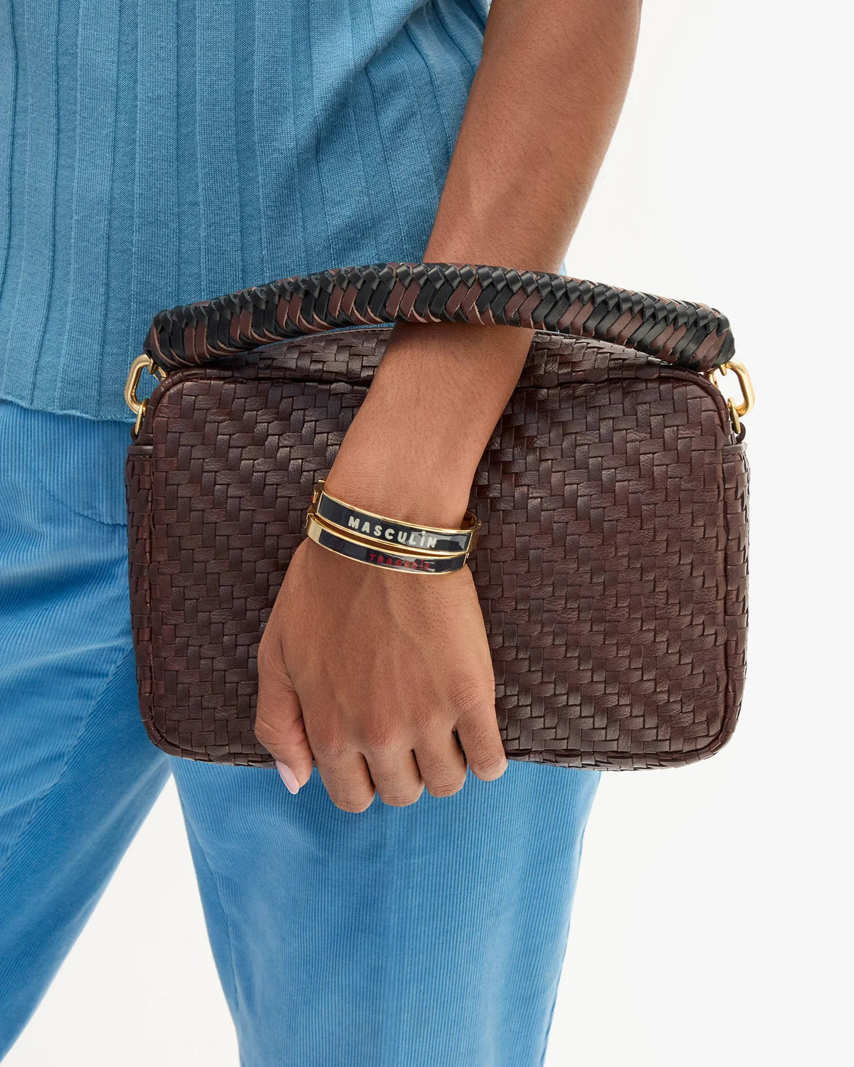 A person wearing a blue ribbed top and blue pants holds a dark brown woven clutch bag with a braided handle and gold-tone hardware. 