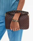 A person wearing a blue ribbed top and blue pants holds a dark brown woven clutch bag with a braided handle and gold-tone hardware. 