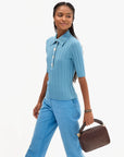 A woman modeling a light blue ribbed short-sleeve button-front top and matching blue trousers, holding a brown woven handbag against a plain light gray studio background.