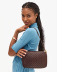A woman with braided hair wearing a light blue ribbed top, carrying a brown woven leather shoulder bag with gold-tone hardware against a plain white background.
