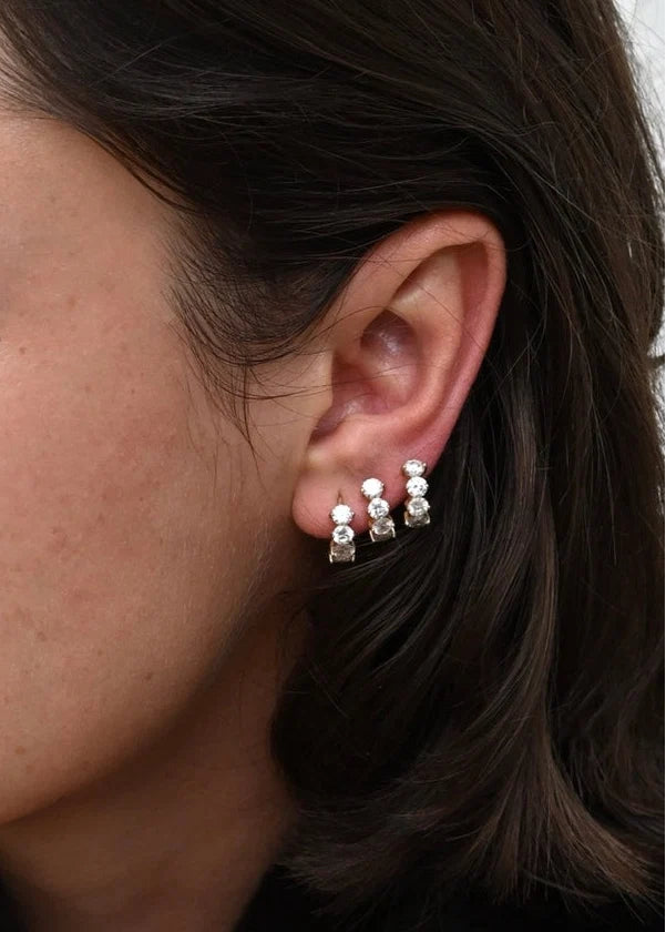 A close-up of a woman's ear wearing three small hoop earrings with clear gemstones, shown against her dark hair and skin.