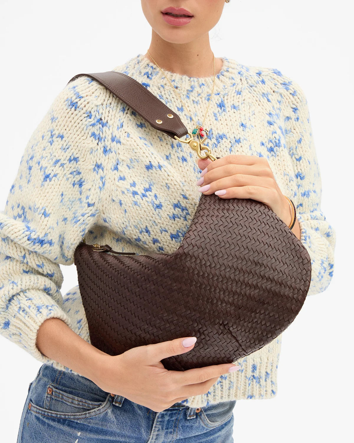 A woman wearing a cream knit sweater with blue floral patterns and blue jeans holds a dark brown woven shoulder bag with a wide brown strap and gold-tone hardware against a plain white background.